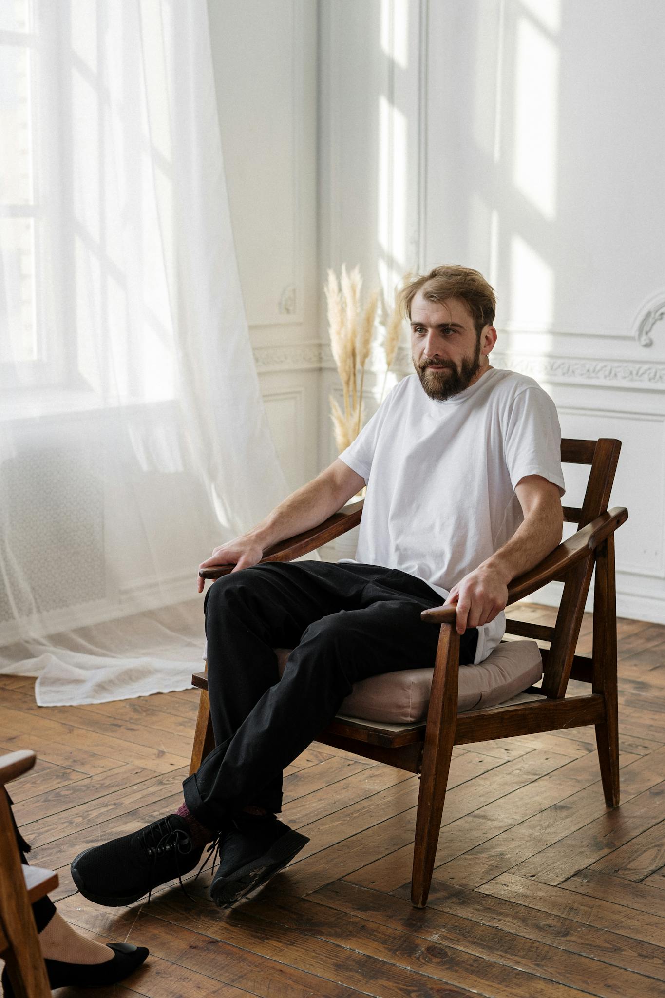 A bearded man in a white t-shirt sits thoughtfully in a chair in a sunlit room.